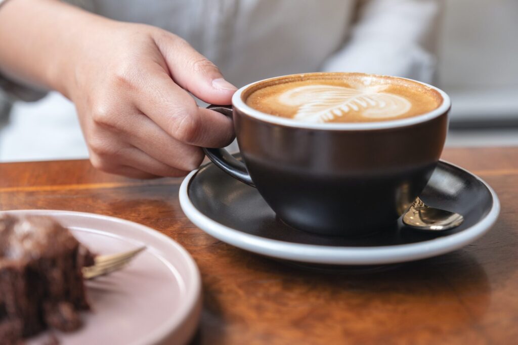 Closeup image of a woman's hand holding and drinking hot latte coffee with brownie cake on the table